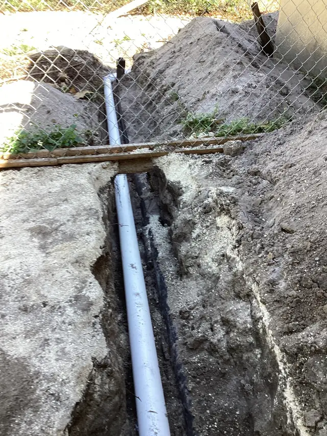 New white PVC drain line laid in an open trench in sandy soil beside a chain-link fence